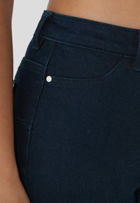 Jeans de mezclilla azul oscuro con una textura suave, cintura alta y costuras de bolsillo frontal visibles. Presenta un detalle de botón plateado.