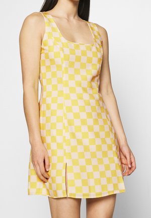 Nainen, joka pukeutunut hihattomaan kelta-valkoiseen ruutukuosiseen mekkoon, jossa on etuhaara, kuva on otettu hartioista reiden keskivaiheille yksivärisellä taustalla.