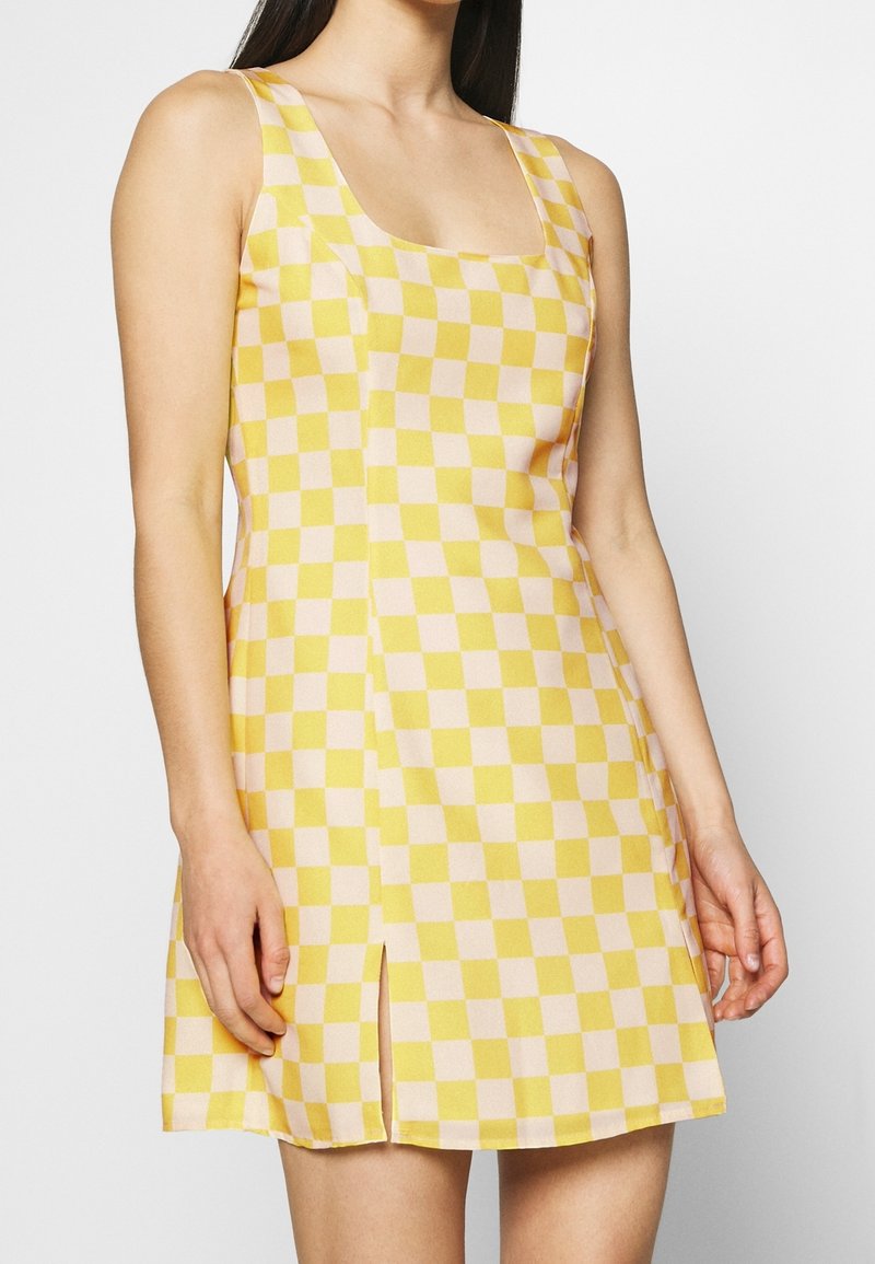 Femme portant une robe sans manches à carreaux jaunes et blancs avec une fente à l'avant, montrée des épaules jusqu'à la mi-cuisse sur un fond uni.