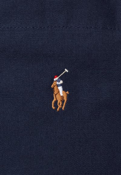 Παίκτης πόλο με ρόπαλο πάνω σε άλογο, κεντημένος σε καφέ, άσπρο, σκούρο μπλε και κόκκινο χρώμα σε φόντο από σκούρο μπλε ύφασμα.