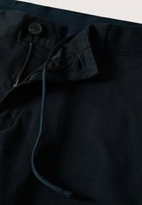 Pantalon bleu marine foncé avec fermeture à bouton et zipper, cordon de serrage, et texture de tissu lisse, avec un détail de ceinture visible.