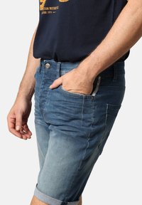 Shorts en denim déclinés dans une gamme de tons de bleu, avec un ourlet roulé, des poches avant et une fermeture à bouton en métal. Texture usée observée.