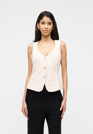 Femme aux longs cheveux noirs portant un gilet crème sans manches boutonné et un pantalon noir, debout devant un fond clair uni.