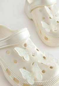 Weiße geformte Clogs mit einer strukturierten Oberfläche, die vier durchsichtige Schmetterlingsdekorationen mit glitzerndem Glanz auf der Oberseite aufweisen.