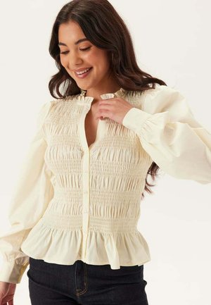 Ung kvinna som ler, iklädd en krämfärgad blus med smock och långa puffärmar med knappar, kombinerad med mörka jeans.