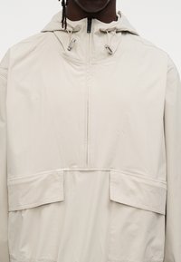 Ανοιχτό καφέ μπουφάν αδιάβροχο με κουκούλα, διαθέτει κλείσιμο με φερμουάρ στο μπροστινό μέρος που φτάνει μέχρι τη μέση, δύο μεγάλες μπροστινές τσέπες και λείο, υφασμένο ύφασμα.