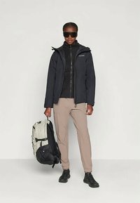 Udendørsjakke i sort, lagdelt over en sort lynlås-overdel, beige bukser og sorte sko. Bærer en tofarvet rygsæk, model med solbriller.