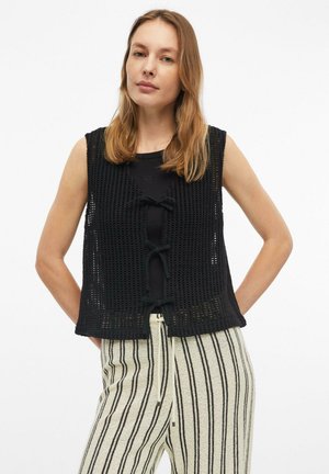 Jonge vrouw die een zwarte mouwloze gehaakte vest draagt, vastgeknoopt met drie strikken over een zwart topje en een witte broek met verticale zwarte strepen.