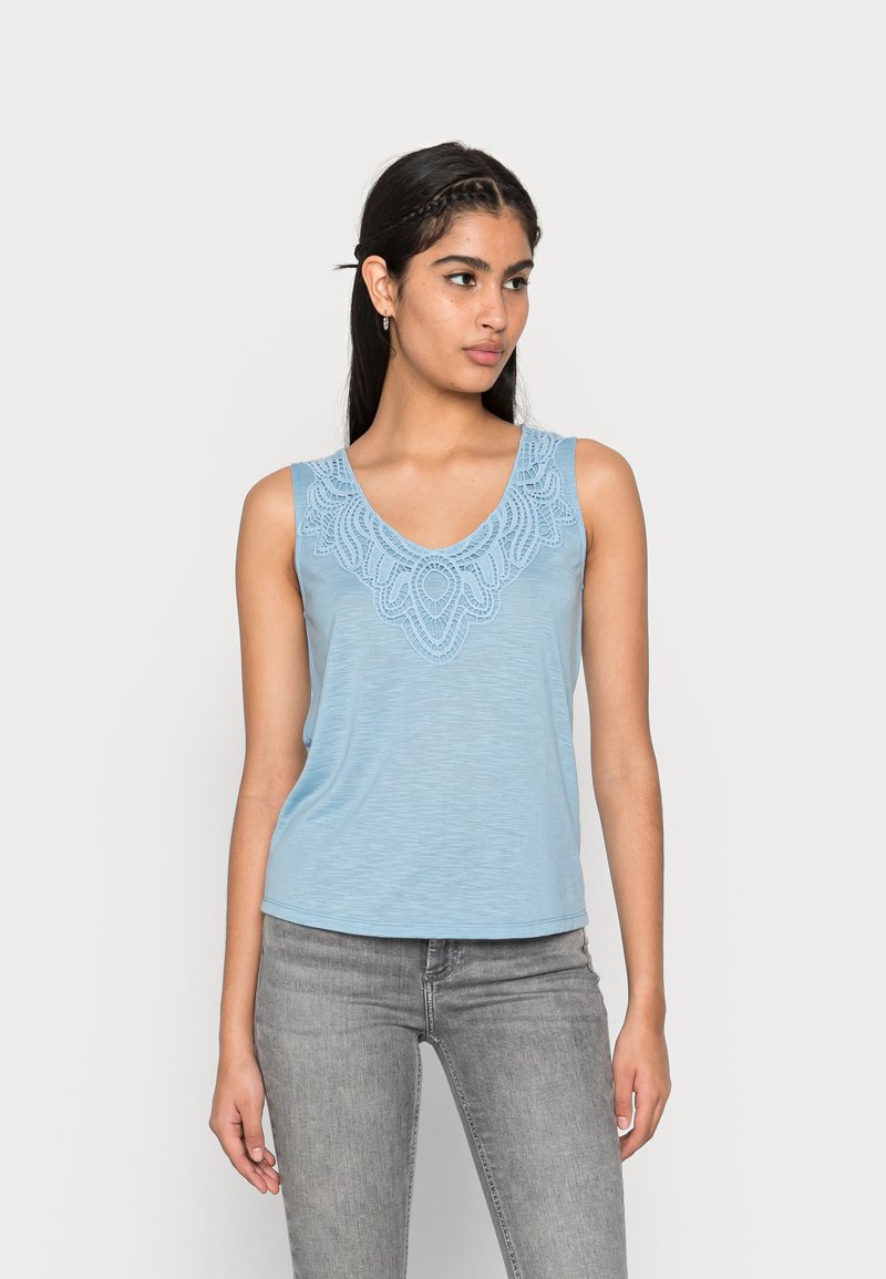 Top sem mangas azul claro com decote em V, apresentando bordados intricados à volta do pescoço. Tecido suave, corte descontraído, combinado com jeans cinzentos.