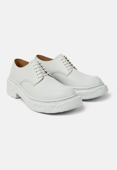 Pari valkoisia nahkaisia nauhallisia kenkiä, joissa paksut kuvioidut pohjat, pyöreät kärjet ja näkyvät ompeleet, yksinkertaisella vaalealla taustalla.