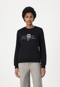 Črna kapuca iz bombažne mešanice, ki vsebuje grafični tisk likovne figure in besedo "KARL." Okrogel ovratnik in rebrasti manšeti s širokim krojem.
