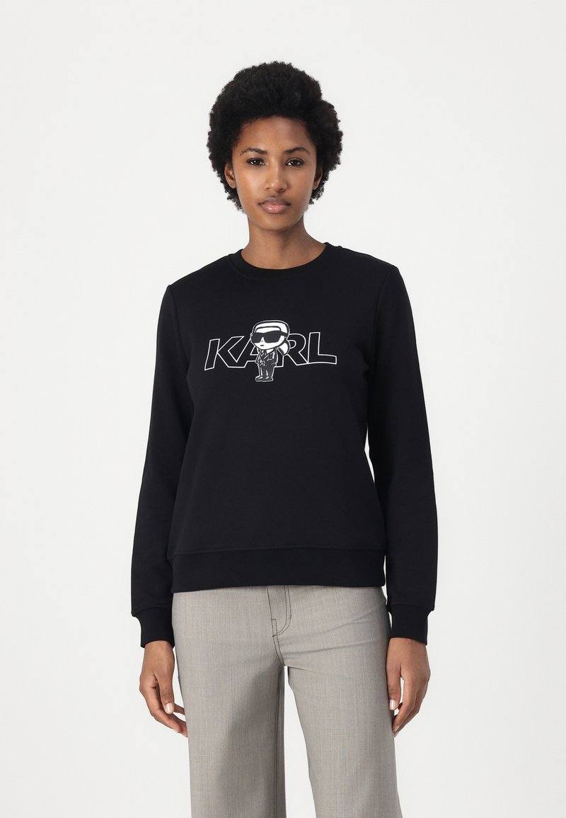 Črna kapuca iz bombažne mešanice, ki vsebuje grafični tisk likovne figure in besedo "KARL." Okrogel ovratnik in rebrasti manšeti s širokim krojem.
