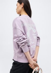 Γυναίκα που φοράει λιλά φούτερ με αφηρημένο πρόσωπο και τη φράση «Women for Women», τα χέρια πίσω από την πλάτη κρατώντας το λουρί μιας μπεζ τσάντας.