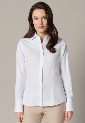 Kobieta o długich brązowych włosach, ubrana w dopasowaną białą koszulę zapinaną na guziki i beżowe spodnie, stojąca na jednolitym jasnym tle.