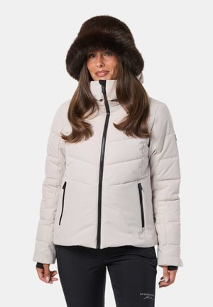 Femme portant une veste d'hiver légère matelassée avec des fermetures éclair noires et un chapeau en fourrure marron foncé, debout devant un fond uni.