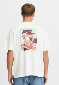 White short-sleeve t-shirt featuring a colorful printed design on the back, depicting a table setting with drinks and people.
