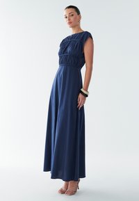 Tamsiai mėlyna maksi suknelė su prigludusiu liemeniu, trumpomis rankovėmis ir surinktomis detalėmis. Lengva medžiaga gražiai krenta iki žemės.