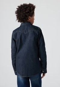 Mørk denimskjorte med lange ermer, spiss krage, synlige sømdetaljer og buet kant. Bakfra vises yoke-design.