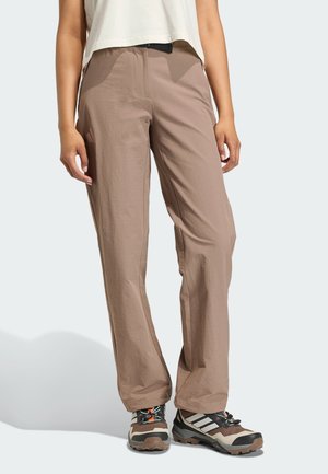 Osoba nosząca beżowe spodnie outdoorowe i brązowe buty trekkingowe Adidas, stojąca na gładkim jasnym tle.