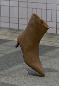 Bottine en daim marron avec bout pointu et talon fin. Dotée d'une fermeture éclair à l'arrière pour un enfilage facile. Surface douce et texturée.