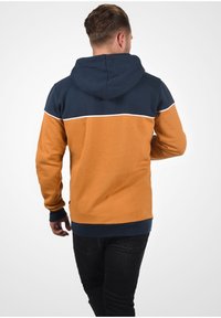 Kapuzensweatshirt mit navy-blauem Oberteil und orangefarbigem Unterteil, versehen mit einem weißen horizontalen Streifen, aus einem weichen, strukturierten Stoff gefertigt.