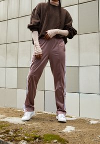 Brązowy, oversize'owy sweatshirt z polaru z prążkowanymi mankietami, w połączeniu z różowymi spodniami dresowymi z satyny z białymi bocznymi paskami, noszony z białymi sneakersami.