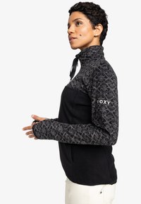 Veste en polaire avec un col montant, dotée d'un corps noir et de manches grises à motifs. Comprend des trous pour les pouces et une fermeture éclair au niveau du cou.