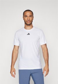 Bela atletska majica s kratkimi rokavi, narejena iz lahkotnega materiala, z okroglim izrezom in črnim Adidasovim logotipom na prsih.