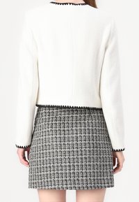 Kvinna iförd en vit texturerad jacka med svarta vågiga kanter över en svart och vit rutig mini kjol, sedd bakifrån.