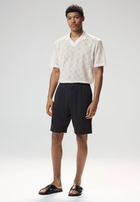 Polo à manches courtes tricoté de couleur crème avec un motif en losanges, associé à un short noir sur mesure et des sandales plates noires, posé sur un fond neutre.