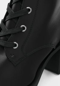 Gros plan sur une botte noire en cuir à lacets avec œillets métalliques, talon épais et coutures apparentes sur une surface en cuir lisse.