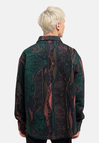 Langermig shirt in donker groen en zwarte stof met abstracte patronen in rood en teal, met een traditionele kraag en een relaxte pasvorm.
