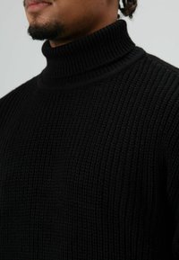 Man draagt een zwarte geribbelde, gebreide coltrui, gefotografeerd van kin tot borst tegen een effen achtergrond.