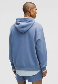 Niebieska bluza z kapturem wykonana z miękkiego materiału, o luźnym kroju, z długimi rękawami oraz kangurzą kieszenią. Warstwowa z jasnoniebieską koszulą pod spodem.