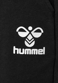 Logo d'abeille stylisé blanc au-dessus du texte "hummel" sur un tissu tricoté noir, centré et clairement visible.