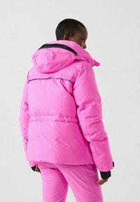 Persona che indossa una giacca invernale imbottita di colore rosa acceso e pantaloni abbinati, di spalle e che guarda verso destra su uno sfondo semplice.