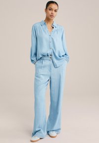 Camisa de mezclilla azul claro con botones en el frente, corte relajado y textura suave, combinada con pantalones de pierna ancha a juego y zapatos casuales.