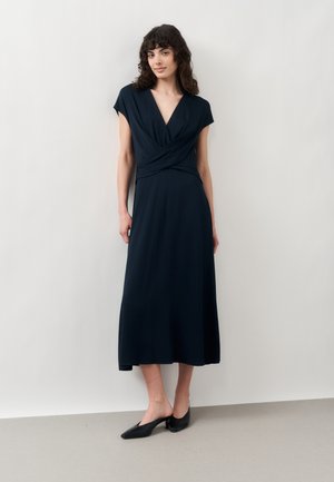 Frau mit dunklem lockigem Haar, die ein schwarzes Midi-Kleid mit V-Ausschnitt und schwarze flache Schuhe trägt, steht vor einem schlichten hellen Hintergrund.