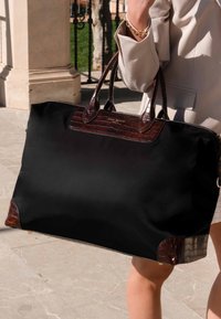 Sac fourre-tout en tissu noir avec des accents en cuir embossé crocodile marron, double anses et fermeture éclair ; texture lisse et forme structurée.