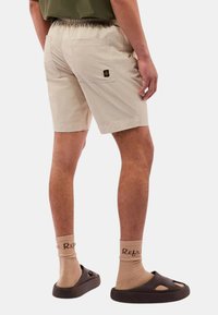 Pantaloni beige con vita elastica, tasche laterali e posteriori, e una piccola etichetta con logo sul retro. Indossati con sandali marroni e calze beige.