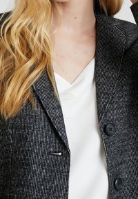 Blazer pied-de-poule in bianco e nero con collo a revers, due bottoni, tasca laterale e indossato sopra una maglia bianca.