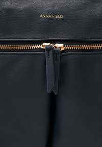 Sac en cuir noir texturé avec une fermeture éclair dorée, doté de deux tirettes et du nom de marque en relief "ANNA FIELD" au-dessus de la fermeture éclair.