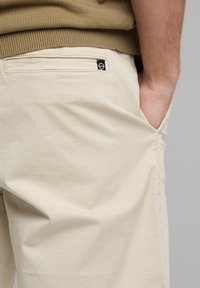 Beige cotton pants with a side pocket and a black logo tag on the back, featuring a smooth, lightweight texture and straight cut design.