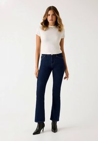 Jean en denim bleu foncé évasé avec une taille haute, associé à un haut blanc ajusté à manches courtes. Le mannequin porte des bottines noires à talons.