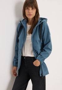 Veste imperméable bleu clair avec capuche, poignets élastiques, poches avant et fermeture éclair. Portée sur une chemise beige et un pantalon noir.