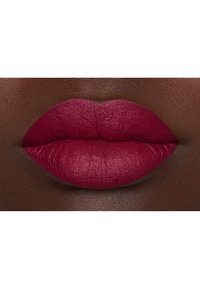 Rouge à lèvres mat couleur baies profondes appliqué sur des lèvres lisses, avec une forme définie et une texture légèrement visible, contrastant contre une peau foncée.