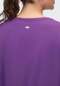 Lila Sportshirt mit rundem Ausschnitt, glatter Textur und einem kleinen Logodetail im Nackenbereich. Leichter und atmungsaktiver Stoff.