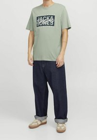 Camiseta de algodón verde claro con un estampado gráfico en azul marino. Pantalones de mezclilla azul oscuro con un corte holgado y puños doblados. Zapatillas beige.