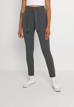 Femme portant un pantalon slim ajusté taille haute à carreaux gris avec une ceinture nouée, un crop top blanc et des baskets blanches montantes.