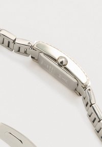 Srebrni sat od nehrđajućeg čelika s pravokutnim kućištem, teksturiranim okvirom i povezanom narukvicom. Na strani ima krunicu za podešavanja.